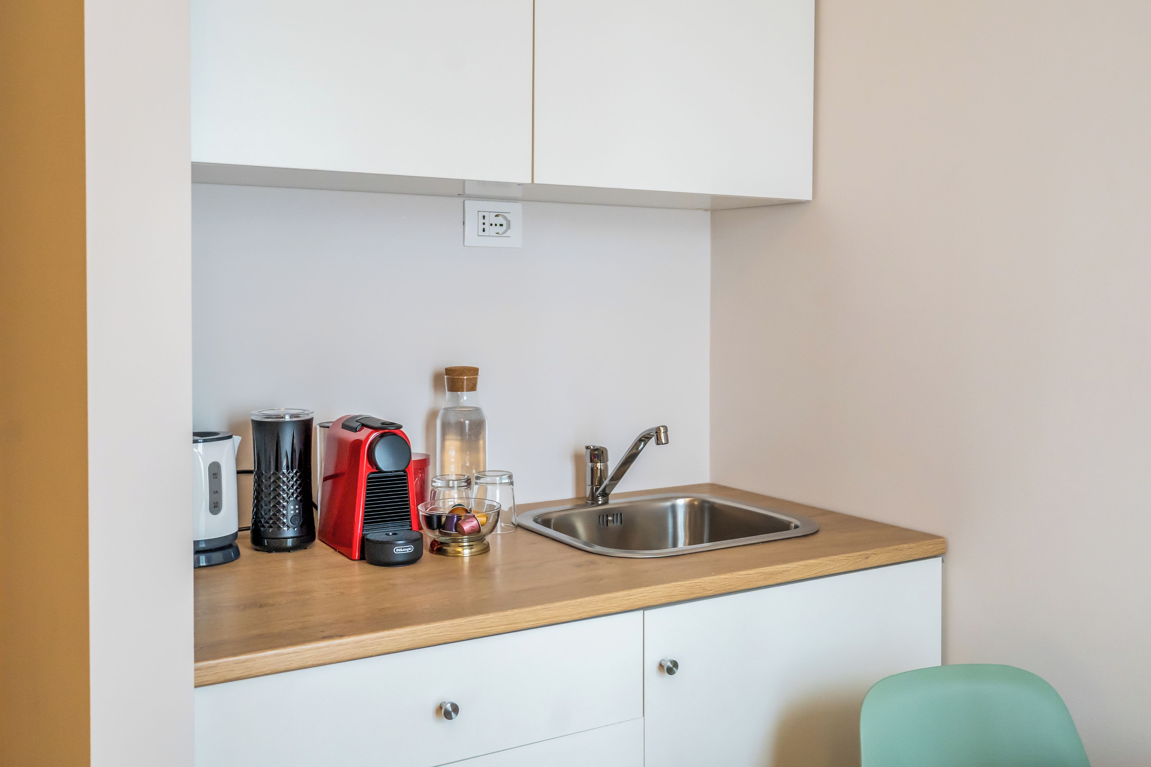 A kitchenette with a stainless steel sink, a red coffee maker, a white kettle, and a glass bottle.