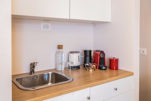 Una kitchenette con un lavamanos de acero inoxidable, una cafetera, un hervidor, un recipiente rojo y un tarro de vidrio.