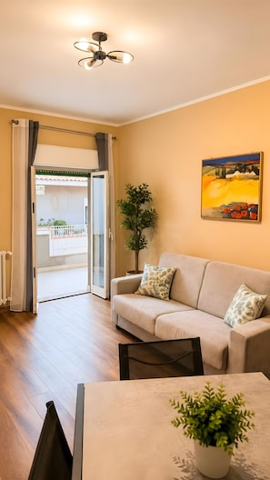 A living room with a sofa, a painting, a potted plant, and a view of the outside.