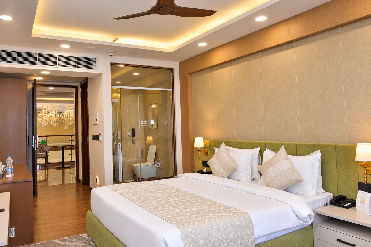 Quarto de hotel com uma cama grande, mesa de cabeceira, escrivaninha, cadeira, chuveiro com divisória de vidro e lustre.