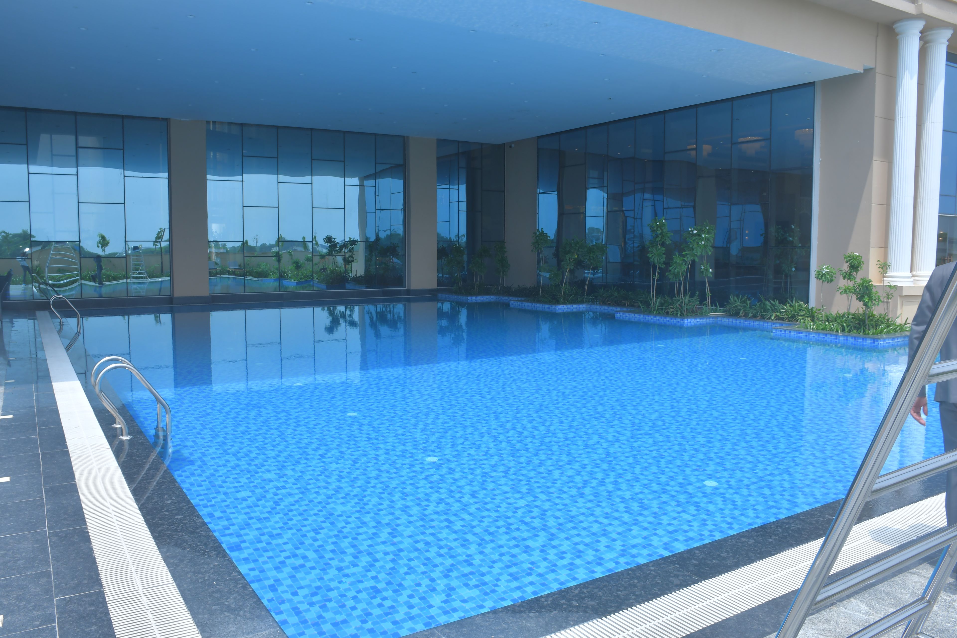 A swimming pool with clear blue water, surrounded by a tiled deck and glass walls reflecting the sky.
