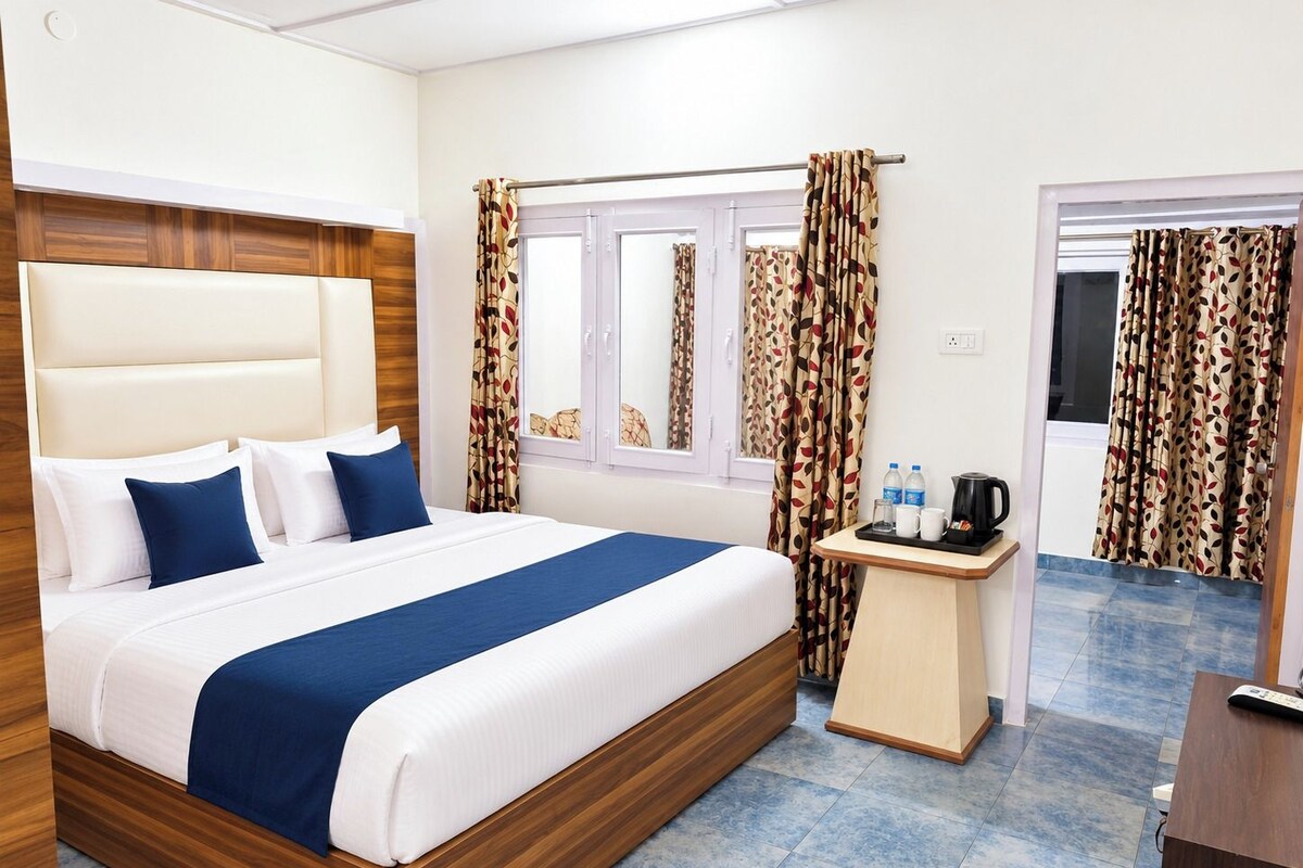 Una habitación de hotel con una cama grande, cabecero de madera, una mesita de noche con una tetera y cortinas con estampado de hojas.