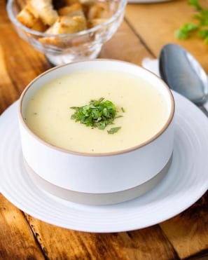 Un tazón de sopa cremosa sobre un plato blanco, con una cuchara, colocado sobre una mesa de madera.