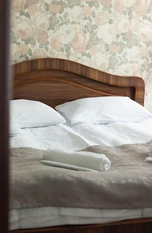 Cama bem arrumada com lençóis brancos e uma toalha enrolada, com papel de parede floral ao fundo.