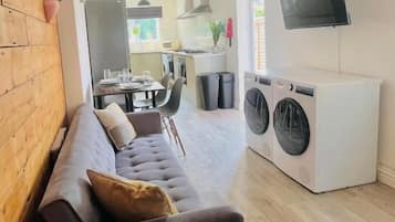 Una sala de estar moderna con un sofá, un televisor instalado en la pared y un área de lavandería con lavadoras.