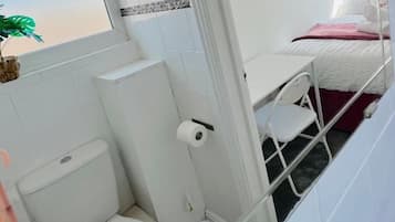 Habitación pequeña con inodoro, lavamanos y cama con colchón blanco y manta rosa.