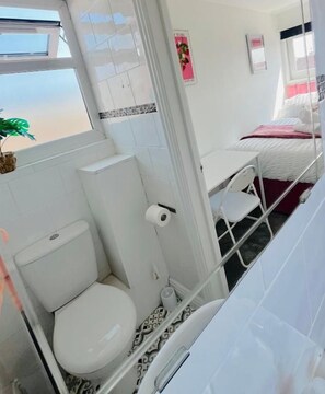 Habitación pequeña con inodoro, lavamanos y cama con colchón blanco y manta rosa.