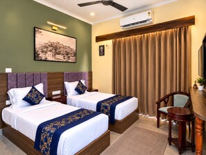 A hotel room with two beds, a desk, a chair, an air conditioning unit, a ceiling fan, and a painting on the wall. - The OG Stays (Jaipur)