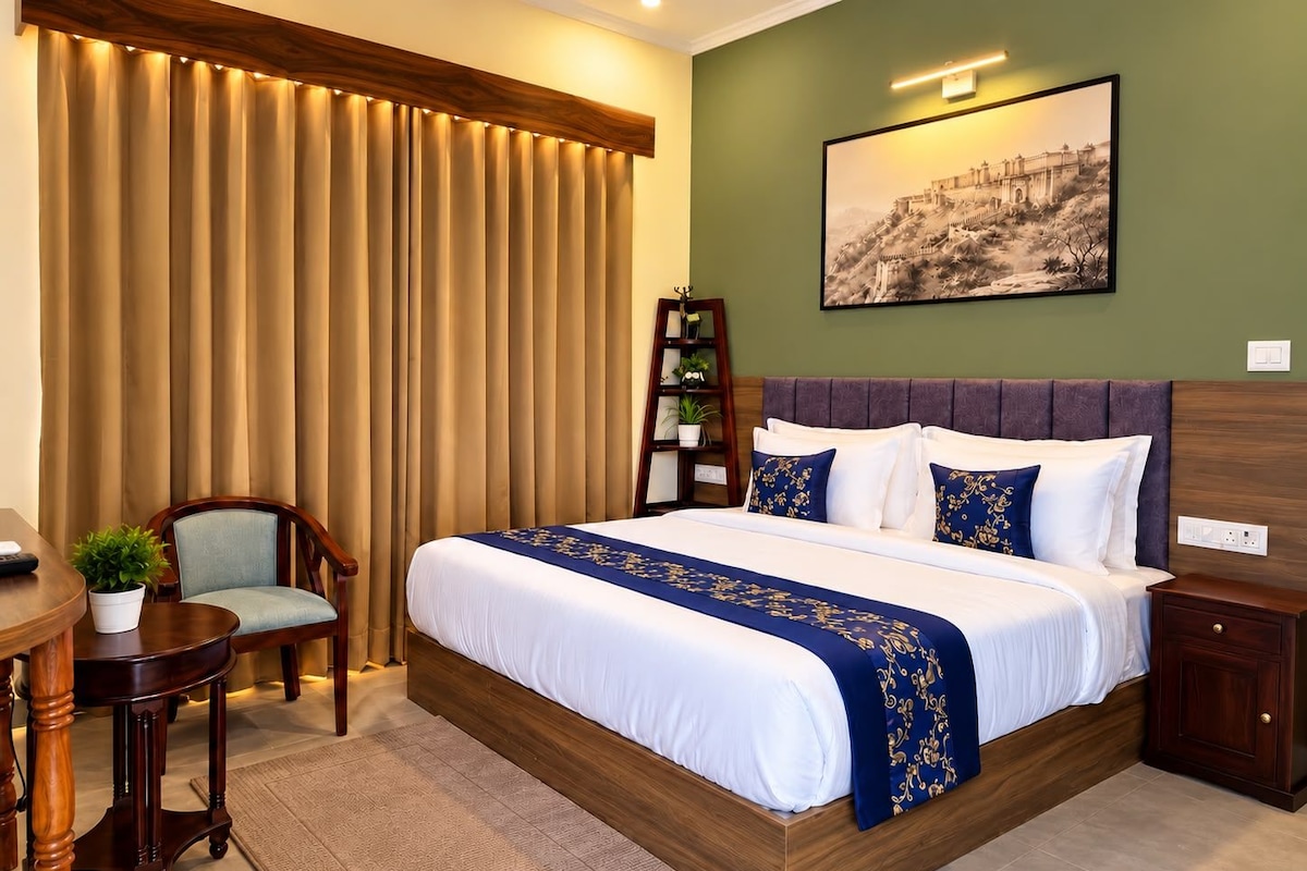 A hotel room with a large bed, a desk, a chair, a small table with a plant, and a bookshelf.