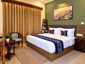 Habitación de hotel con una cama grande, un escritorio, una silla, una pequeña mesa con una planta y una estantería.