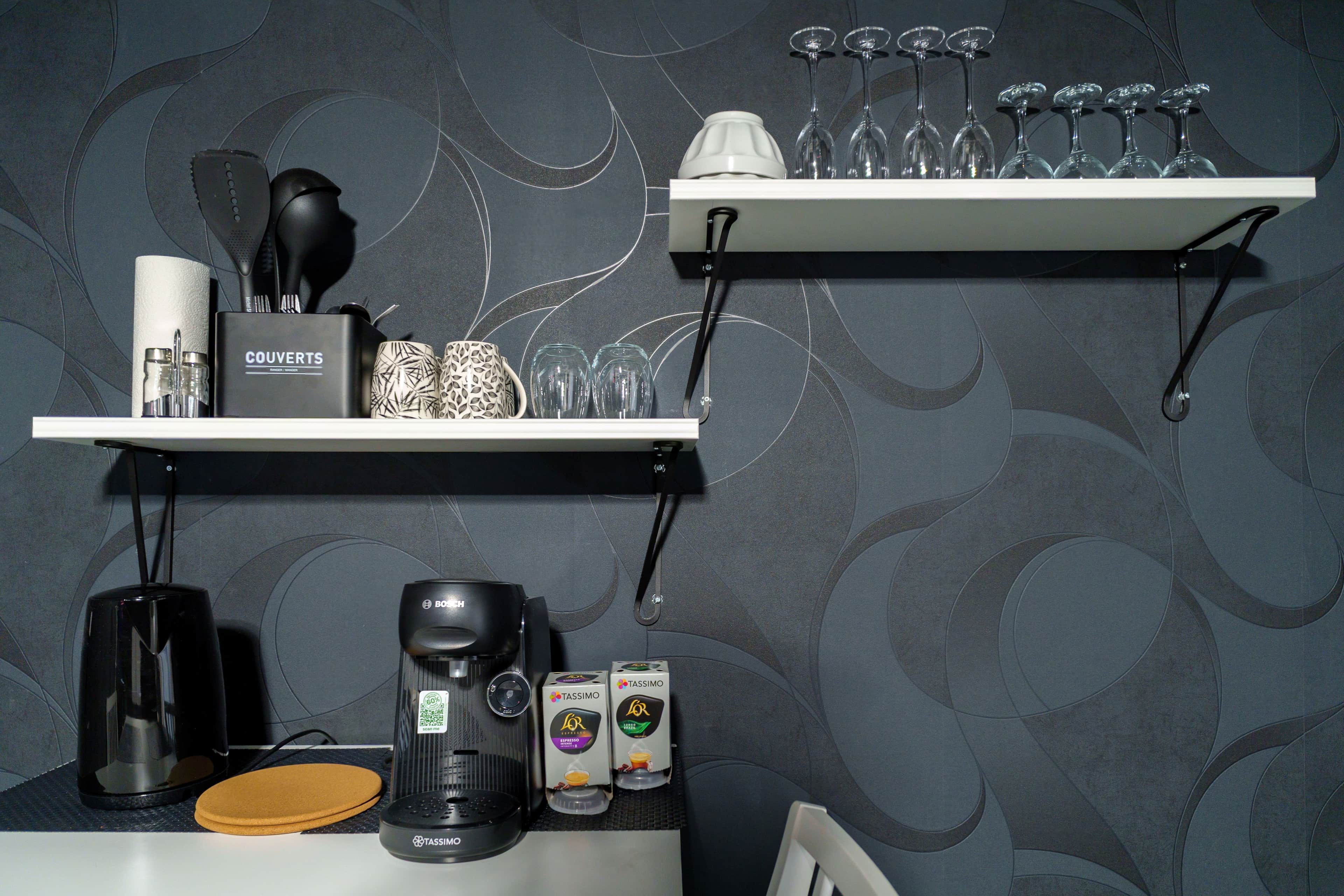Ett köksutrymme med en kaffebryggare, en förpackning med Tassimo-kaffekapslar och ett set vinglas på en hylla.