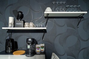 Ett köksutrymme med en kaffebryggare, en förpackning med Tassimo-kaffekapslar och ett set vinglas på en hylla.