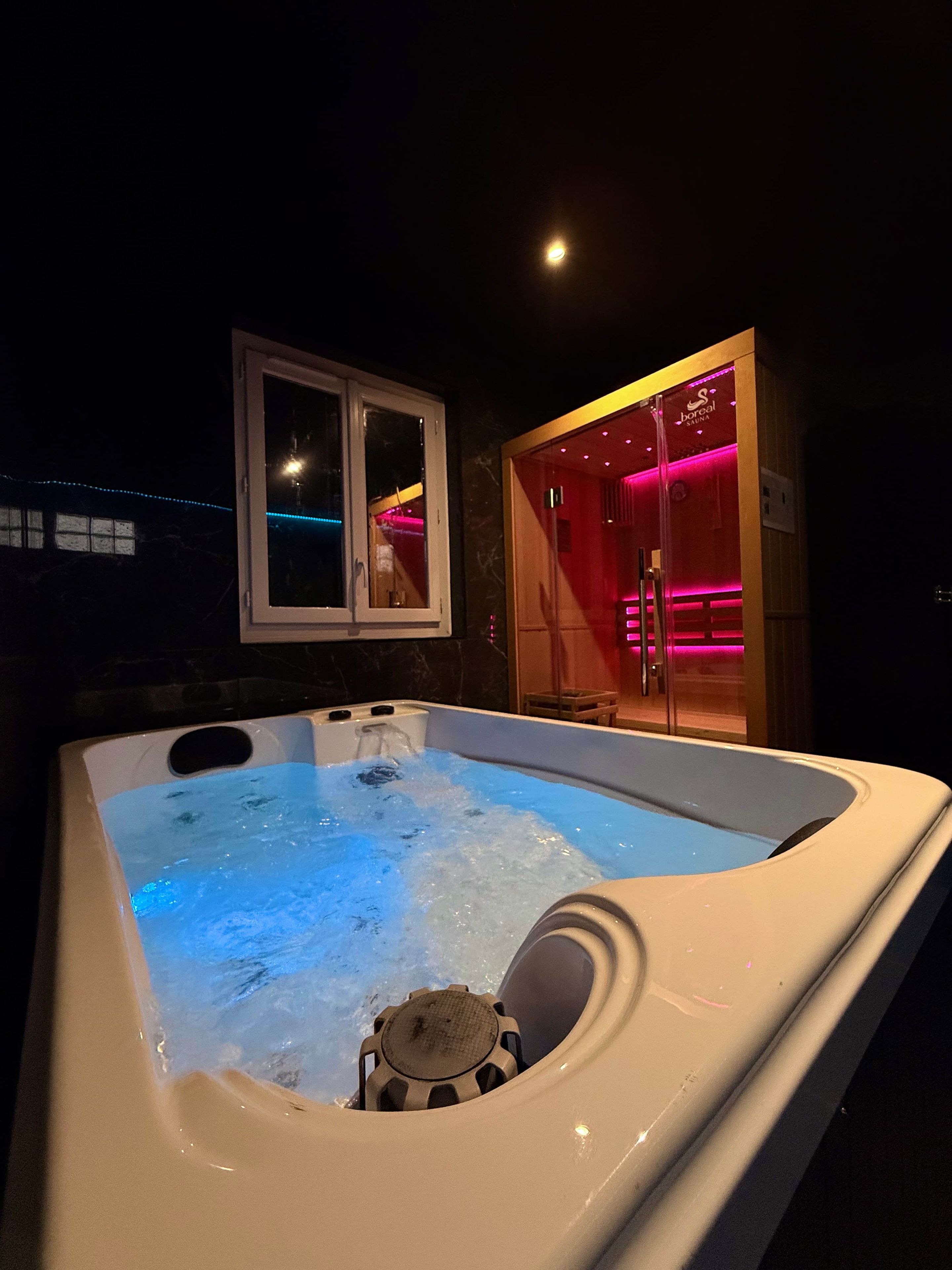 A hot tub with a visible water pump and a wooden structure with red lighting in the background.