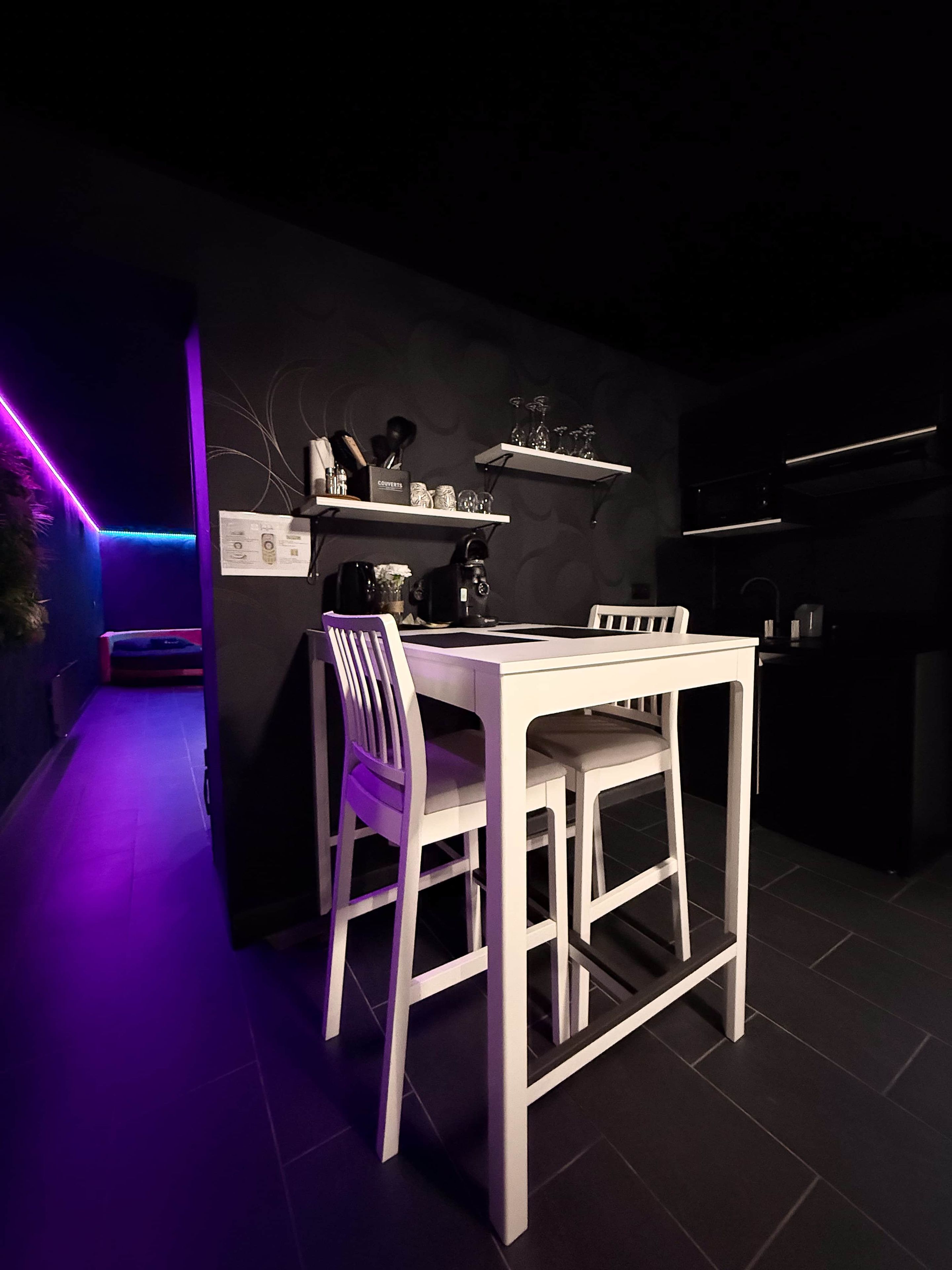 A modern dining area with a white table and chairs, a black wall with shelves holding glasses and a coffee maker, and purple ambient lighting.