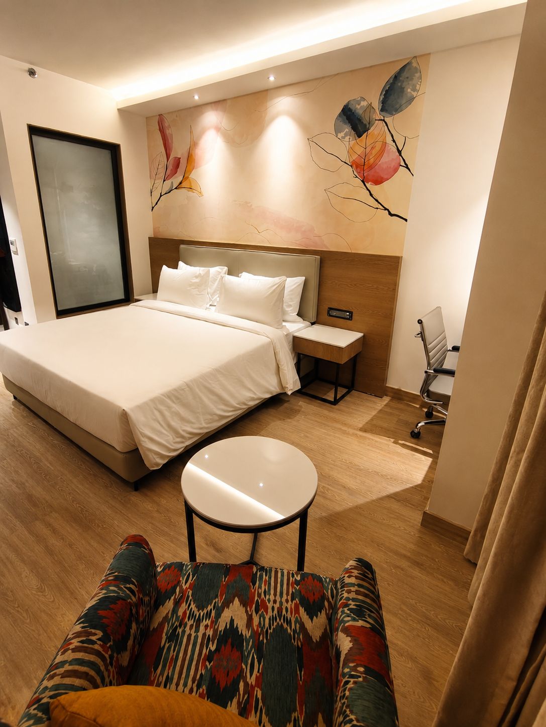 A hotel room with a large bed, bedside tables, a small round table, a colorful sofa, and a wall mural.
