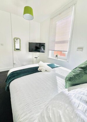 Een hotelkamer met een bed, een televisie, een raam met zonwering en een groene hanglamp.