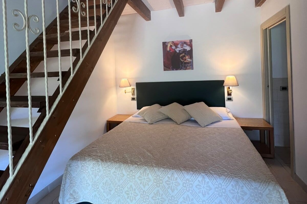 A bedroom with a bed, a headboard, a painting, a staircase, and a nightstand with a lamp.