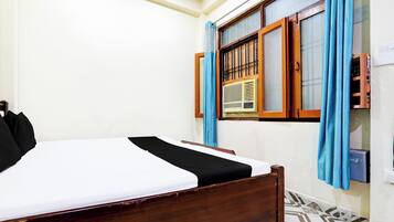 A room with a bed, a window with curtains, a radiator, and a wall-mounted electrical outlet.