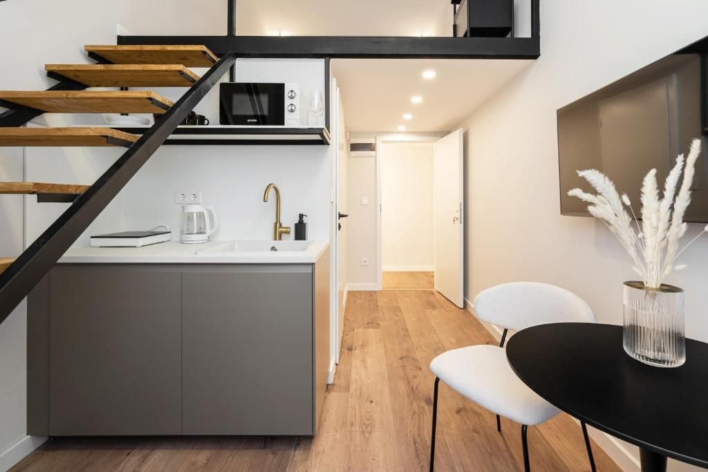 Cocina moderna con microondas integrado, cafetera y pileta. Se observa una escalera de madera que lleva a un rellano. Una mesa negra con un florero de flores blancas es visible.