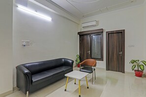 Una habitación con un sofá de cuero negro, una mesita blanca pequeña, una silla marrón, dos plantas en macetas y una puerta de madera con ventanal.