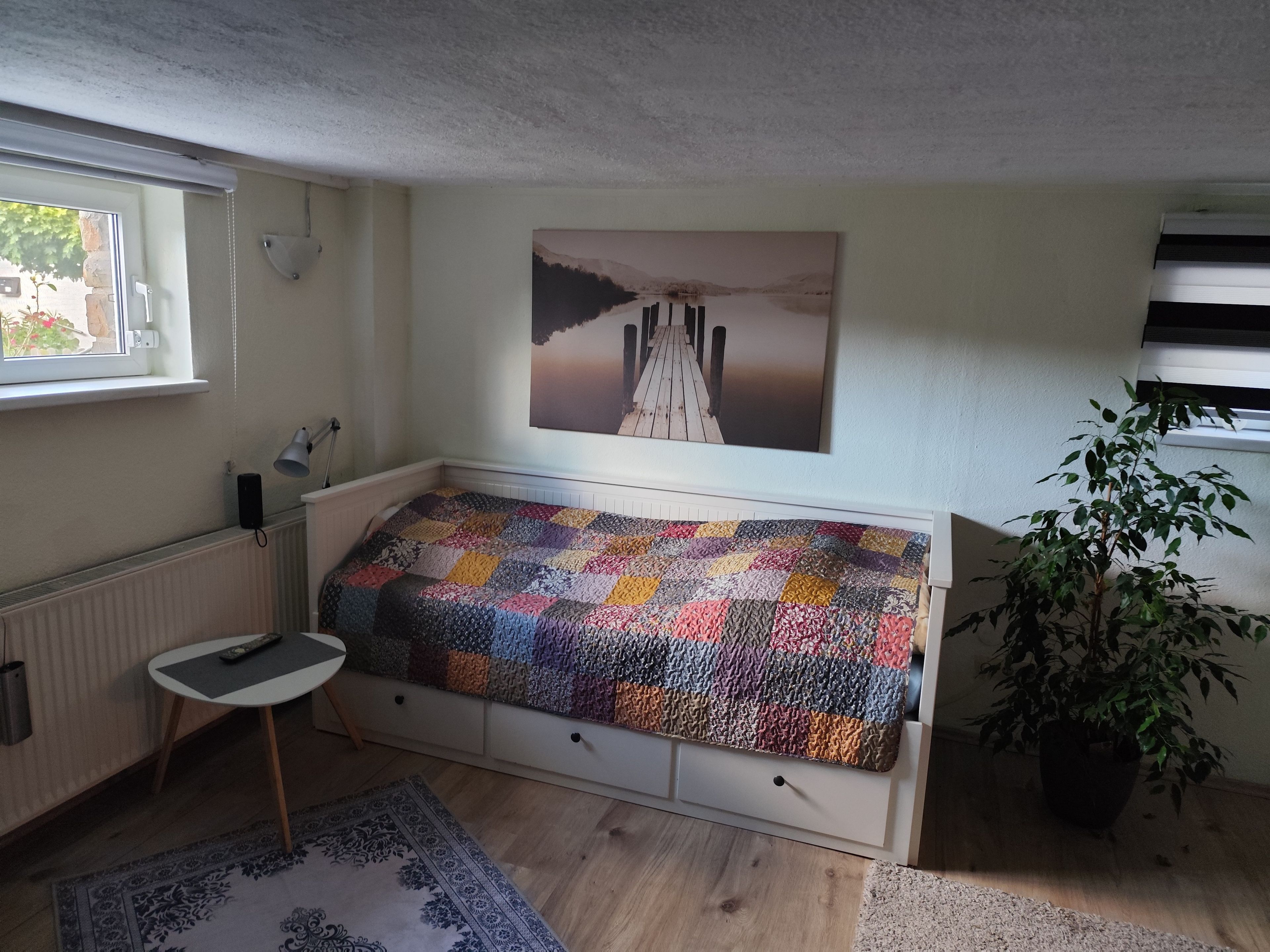 A cozy room with a built-in bed, a radiator, a small table, a potted plant, and a window with a view.