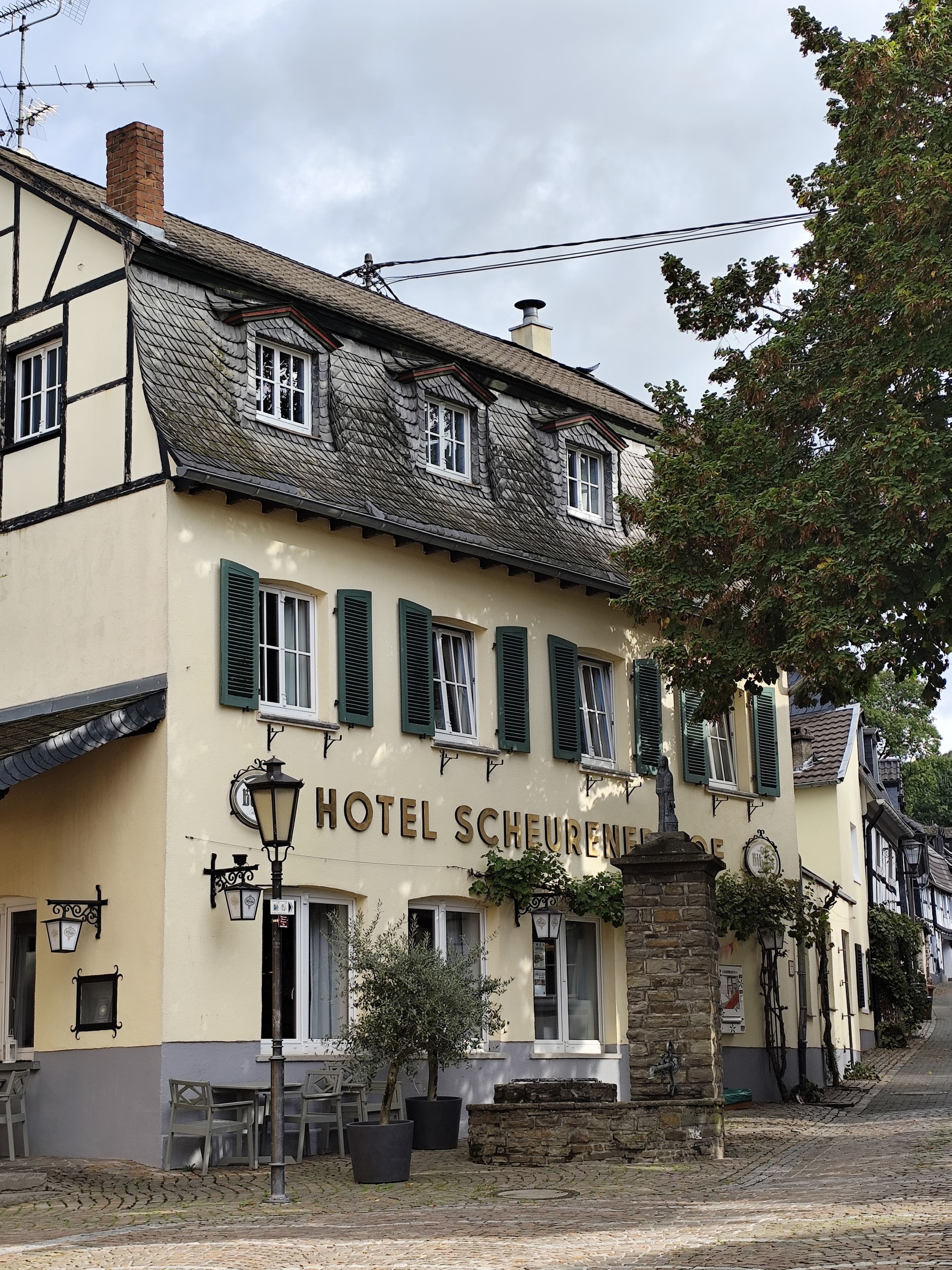 Ein traditionelles Gebäude mit einem Schild, auf dem 'HOTEL SCHEURENSE' steht. Es hat eine Steinfassade, mehrere Fenster mit Fensterläden und eine Kopfsteinpflasterstraße.