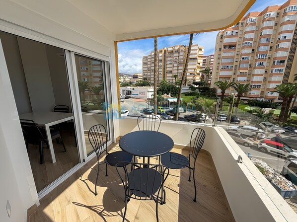 Un balcon avec une table et des chaises, une porte-fenêtre coulissante et une vue sur un quartier résidentiel avec des immeubles et des palmiers.