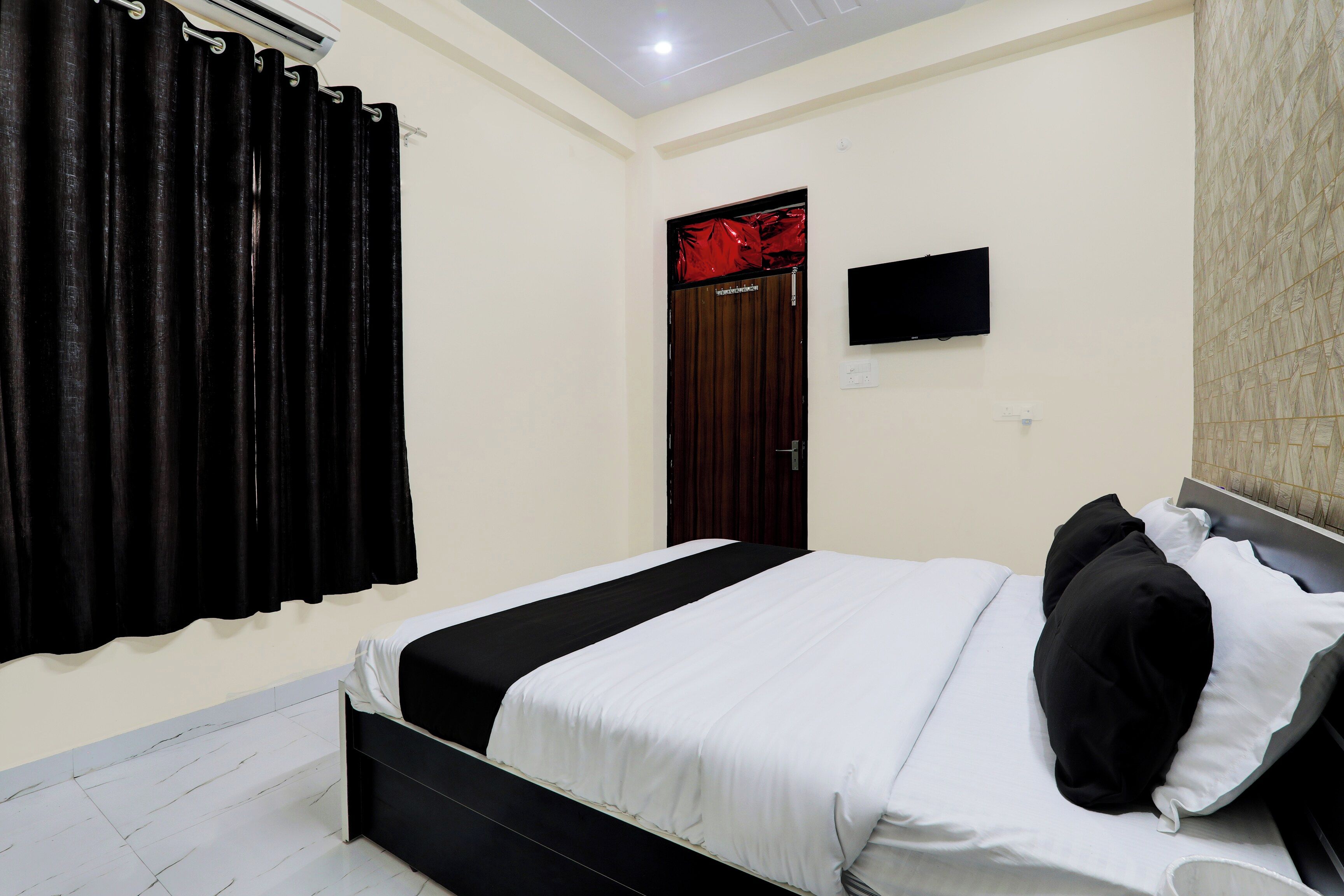 Habitación de hotel con cama, televisor de pantalla plana montado en la pared y ventana con cortinas negras.