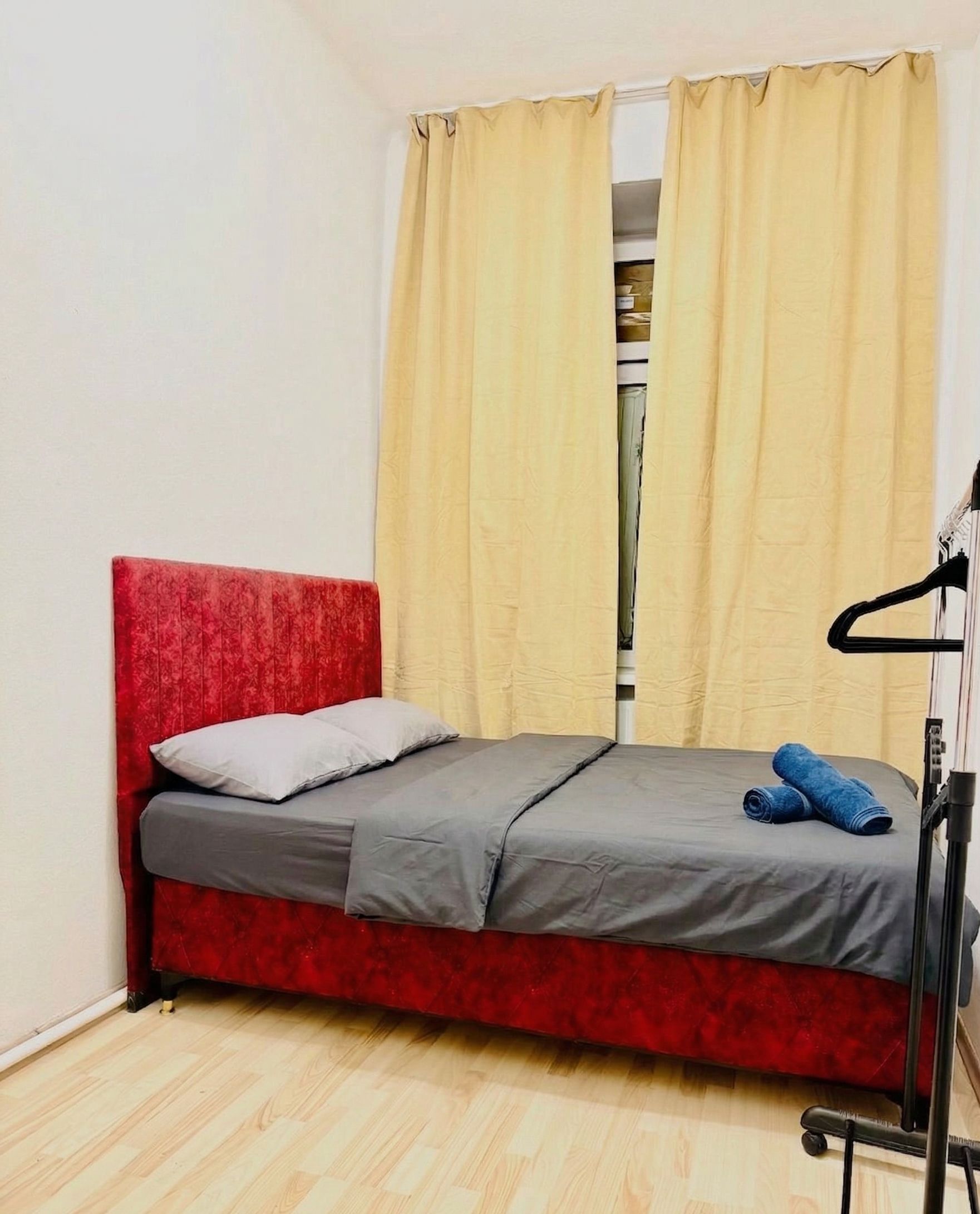 Una habitación pequeña con cama individual, cabecera roja, colchón gris y un par de jeans azules sobre la cama.