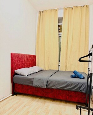 Una habitación pequeña con cama individual, cabecera roja, colchón gris y un par de jeans azules sobre la cama.
