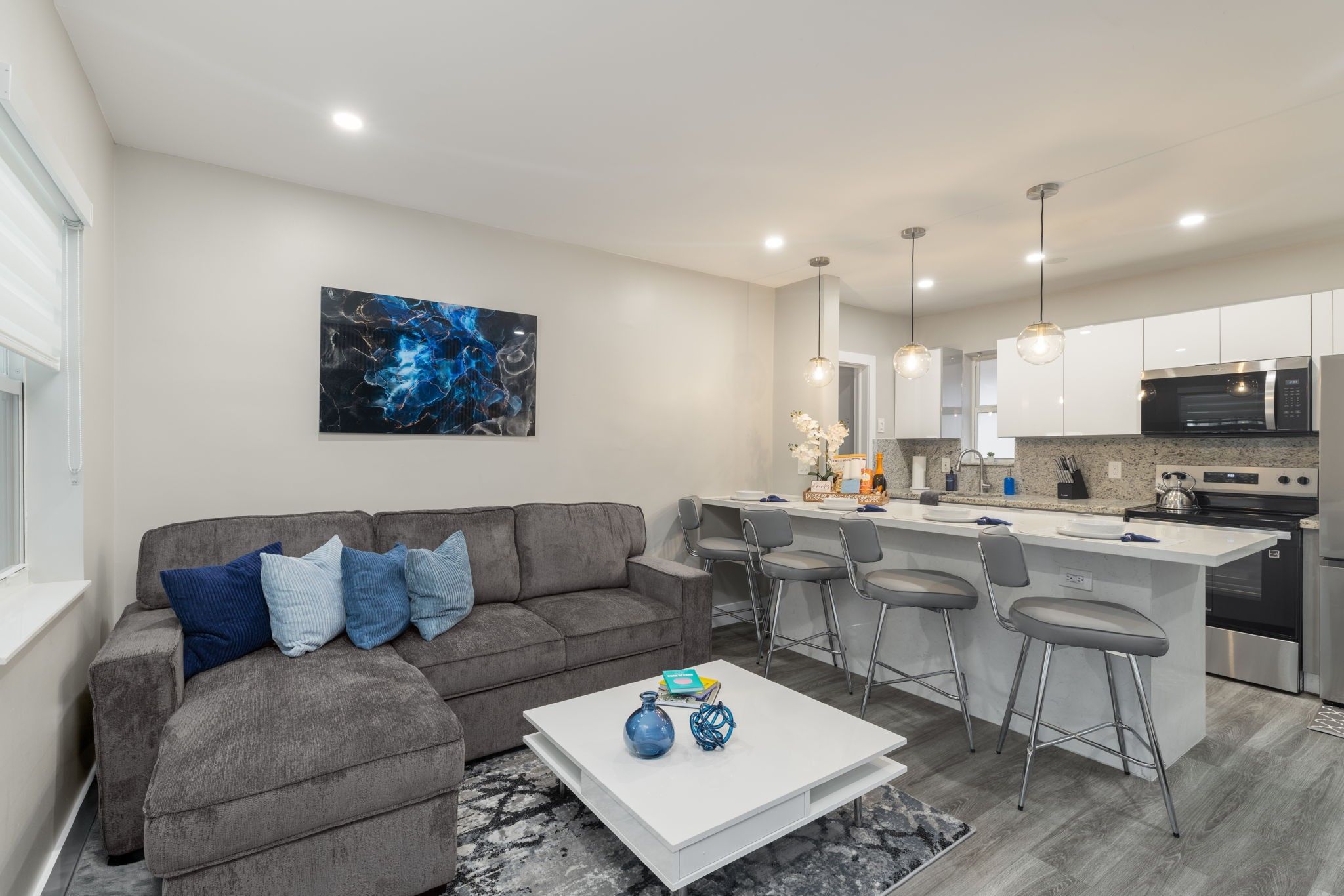 A modern living room with a grey sofa, a coffee table with decorative items, and a kitchen area with a bar stool and a microwave.