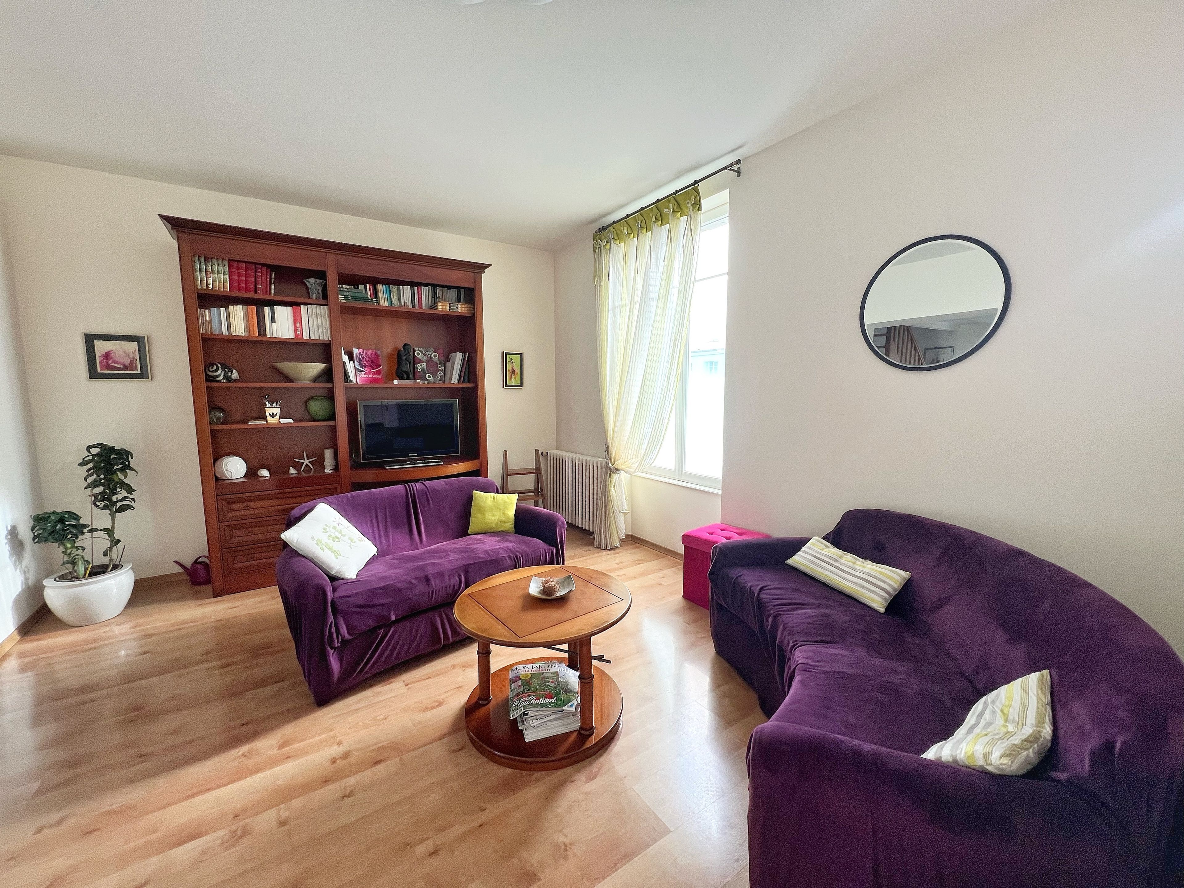 Un salon avec deux canapés violets, une table basse en bois, une bibliothèque, un téléviseur et une fenêtre avec des rideaux.