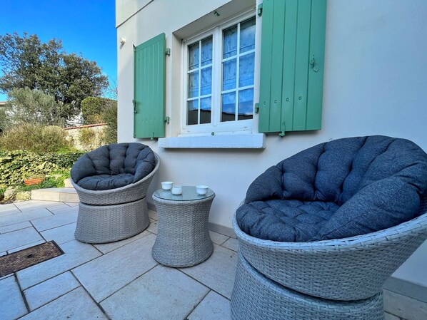 Une terrasse avec deux fauteuils en osier et une petite table, une fenêtre avec des volets verts, et une haie en arrière-plan.