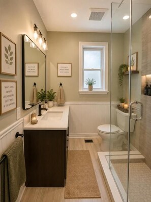 Une salle de bain avec une douche à paroi de verre, un meuble-lavabo à deux vasques, des toilettes et une fenêtre agrémentée d’une plante.