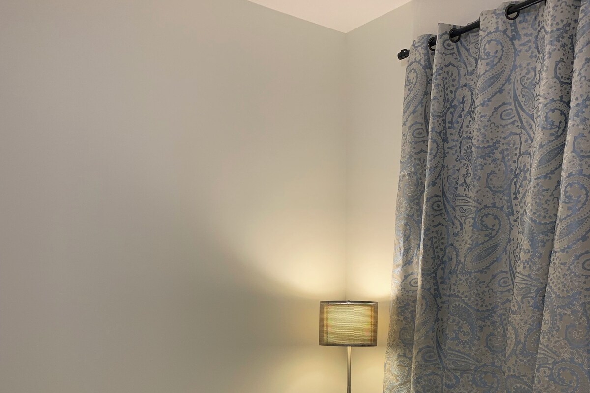 Une chambre à coucher avec un lit, du linge de lit blanc, une table de chevet avec une lampe et un rideau à motifs.
