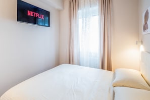 Habitación de hotel con cama, televisor empotrado en la pared mostrando el logotipo de Netflix, ventana con cortinas y lámpara de pared.