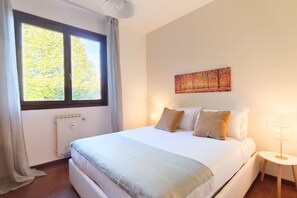 A hotel room with a large bed, a window with curtains, a radiator, a wall-mounted painting, and a bedside table with a lamp.