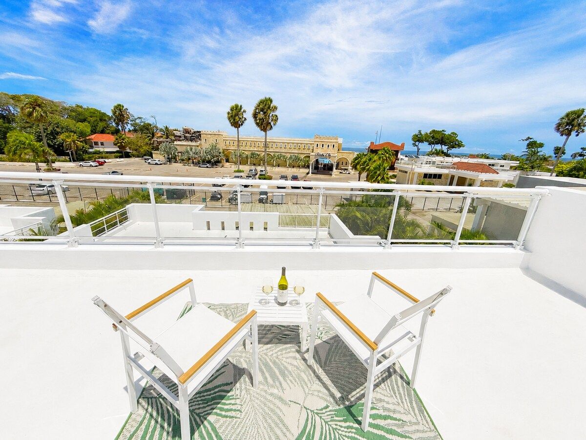 Una terraza en la azotea con una mesa dispuesta para dos personas, con vistas a una zona costera con edificios y palmeras.