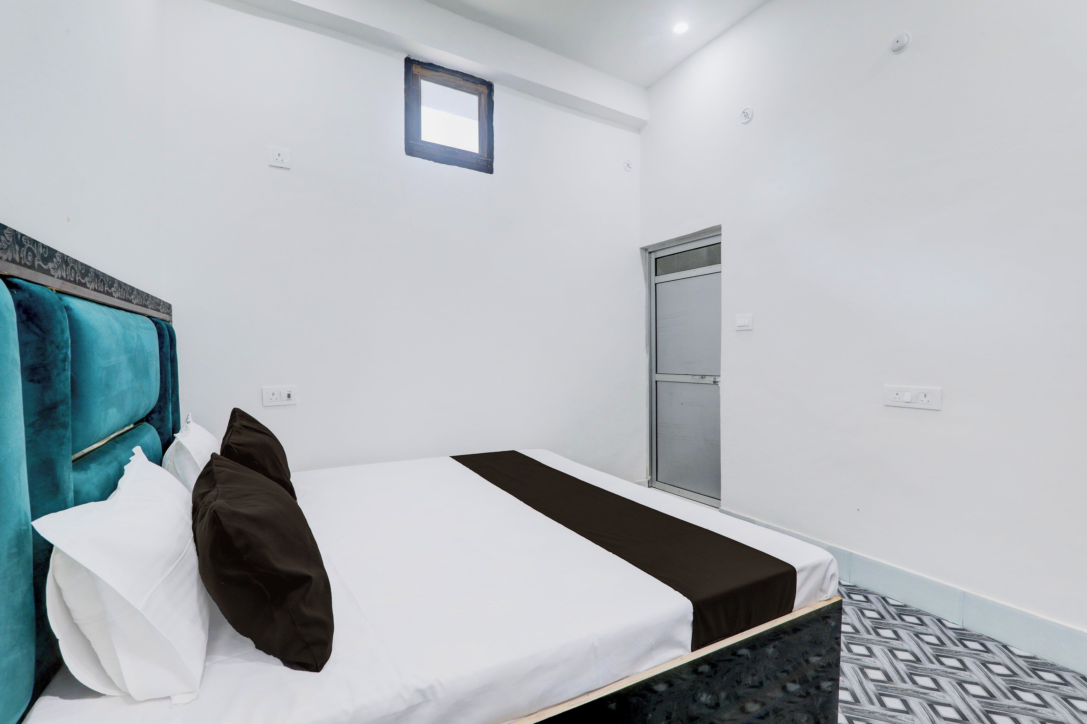 Una cama individual con cabecera color aguamarina, almohadas blancas y marrones, colcha negra y piso estampado.