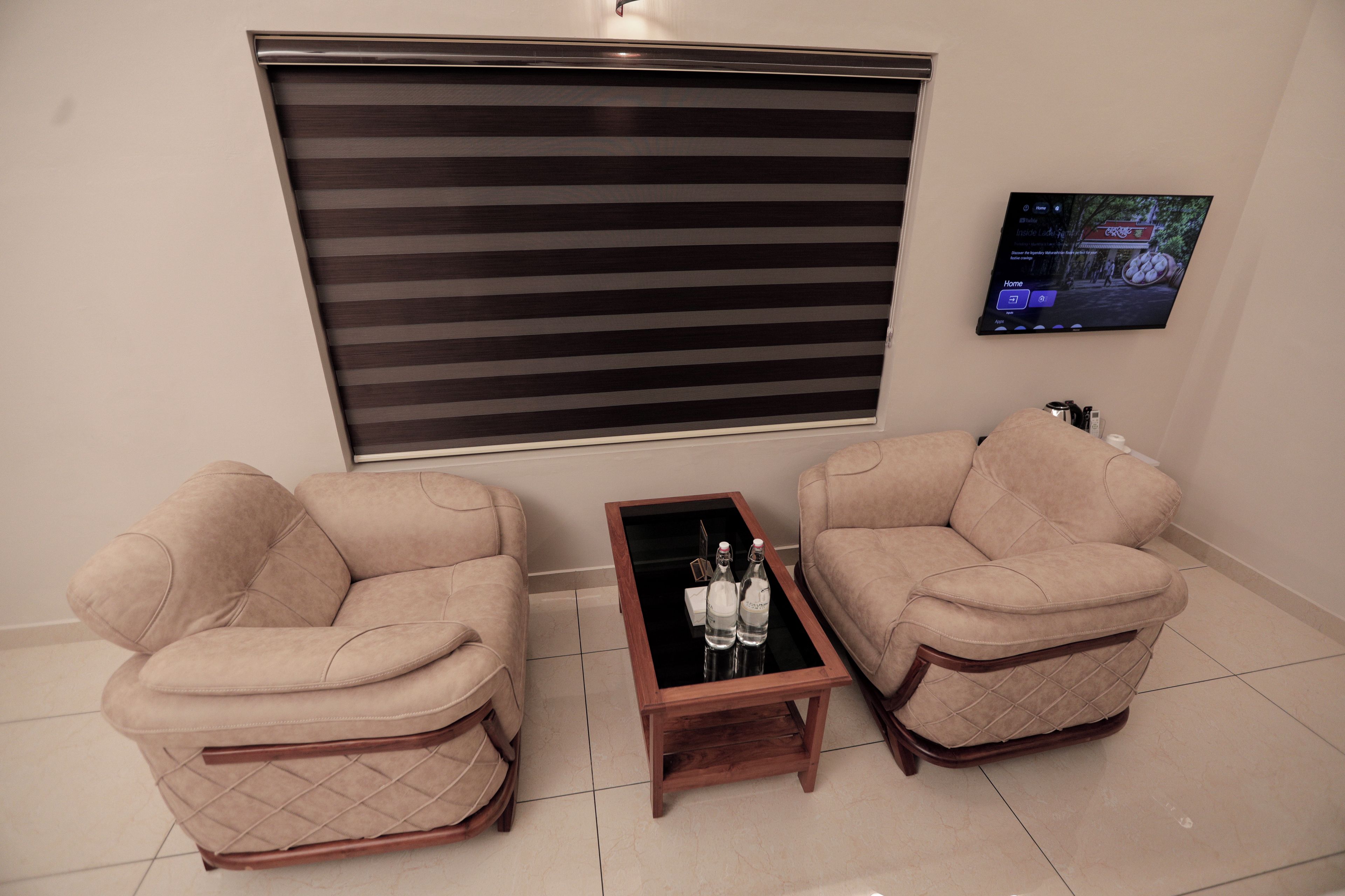 A room with two beige armchairs, a small wooden table with two bottles, and a television mounted on the wall.