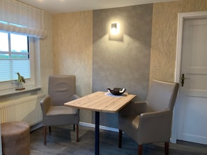 Een kamer met een houten tafel en twee stoelen, een wandlamp, een radiator en een raam met jaloezieën.
