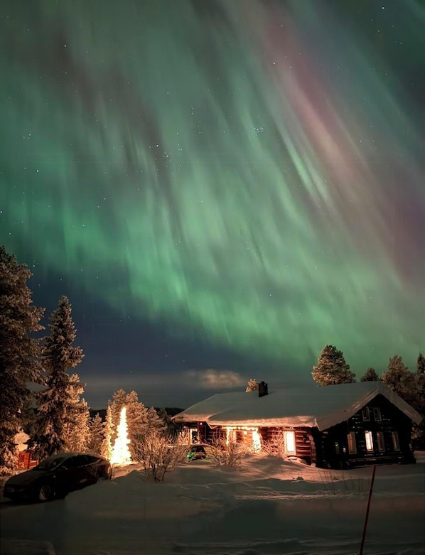 Una cabaña con techo nevado, rodeada de árboles, bajo un cielo nocturno con la aurora boreal.