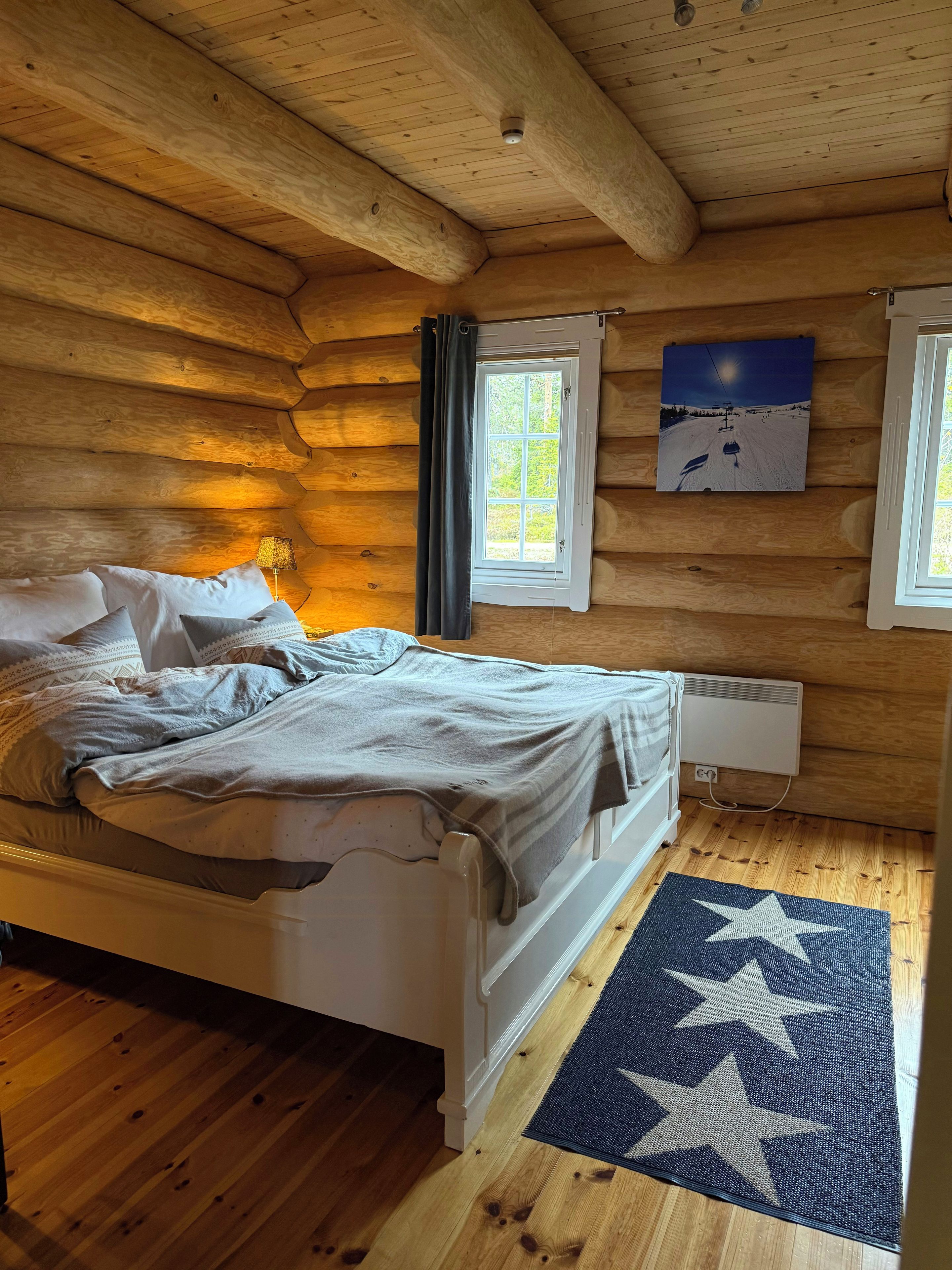 Una habitación de cabaña de madera con una cama, una ventana con cortinas, un radiador y una alfombra con estampado de estrellas.