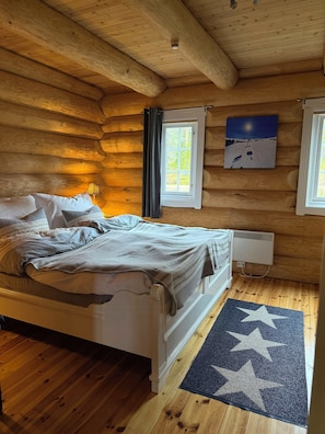 Una habitación de cabaña de madera con una cama, una ventana con cortinas, un radiador y una alfombra con estampado de estrellas.