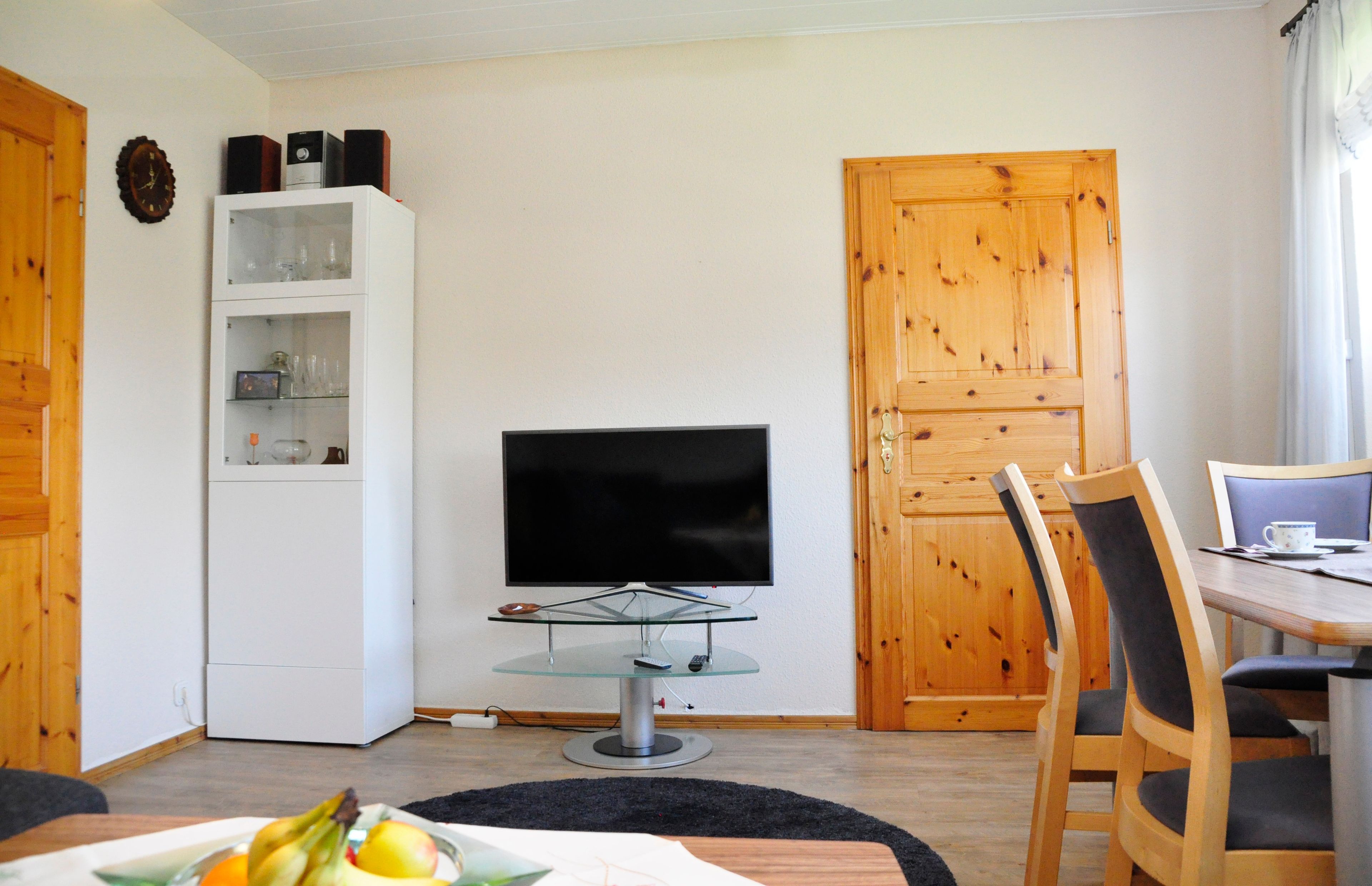 Ein kompakter Wohnbereich mit Fernseher auf einer Glaskonsole, einer Holztür und einem Essbereich mit Tisch und Stühlen.