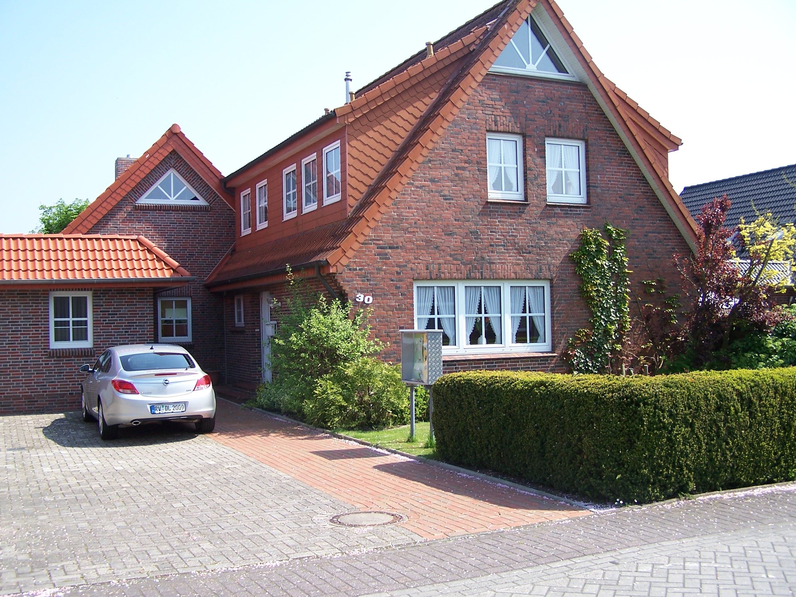 A two-story brick house with a car parked in the driveway, a well-maintained hedge, and a clear sky.
