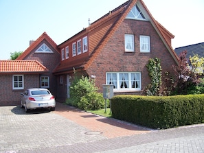 Une maison en briques à deux étages, avec une voiture garée dans l’allée, une haie bien entretenue et un ciel dégagé.