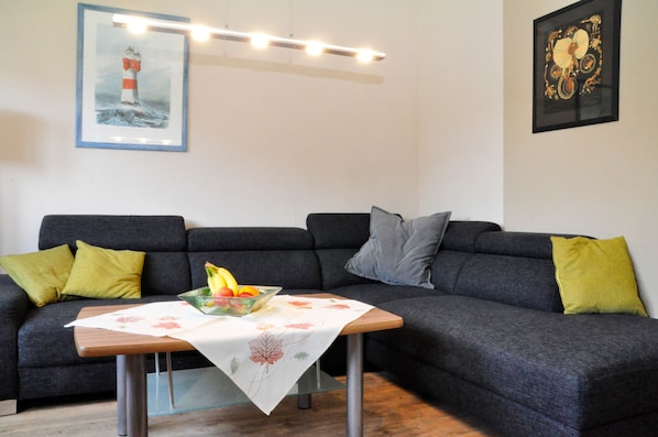 Ein Wohnzimmer mit einem dunkelgrauen Sofa, einem Couchtisch mit Obstschale und einem Gemälde eines Leuchtturms.