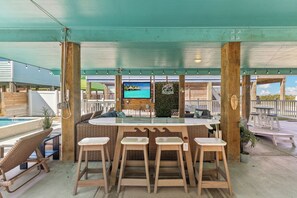 Un espace bar extérieur abrité, avec vue sur la piscine, des tabourets en bois et un écran de télévision.