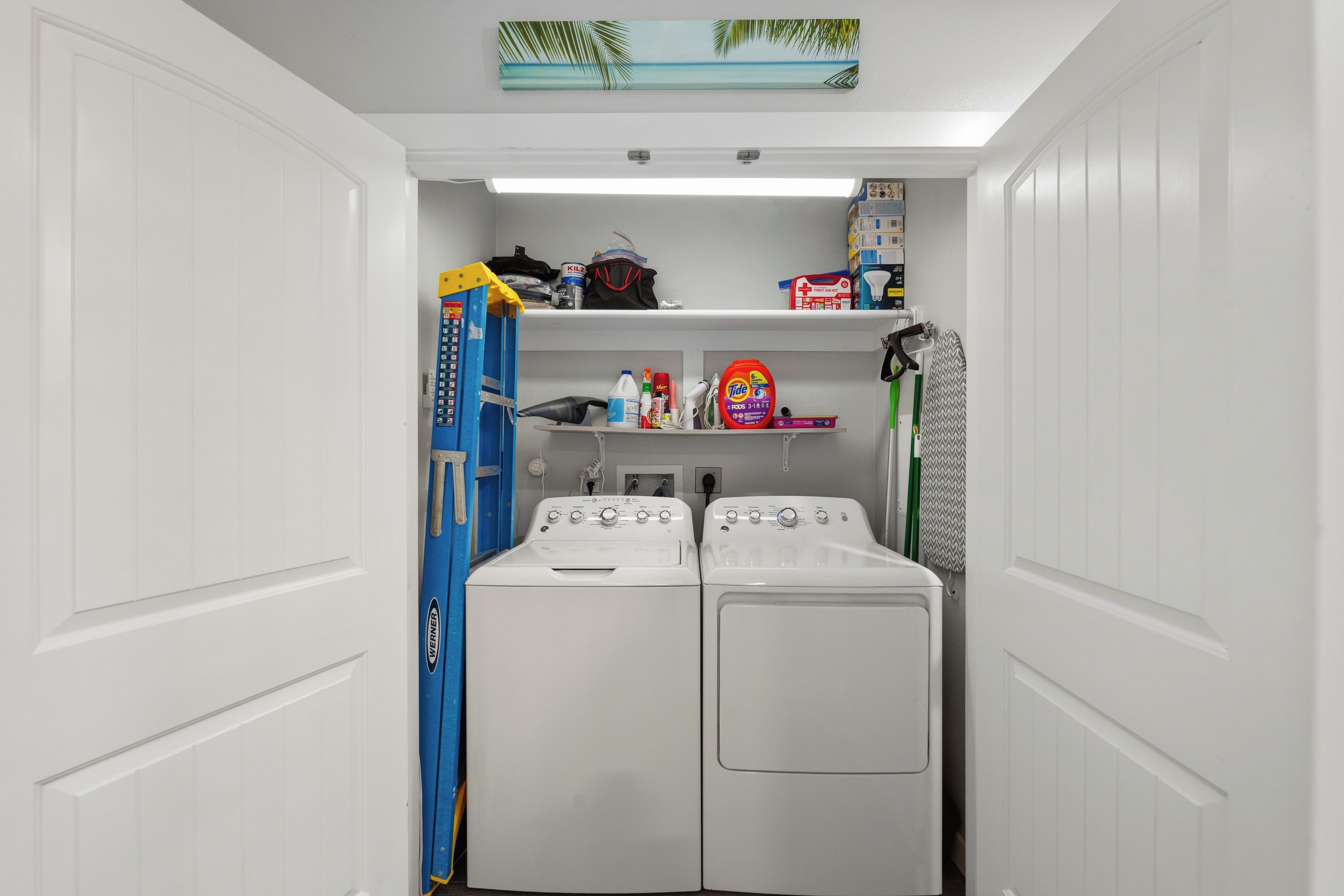 A laundry room with two white front-loading washers, a shelf with cleaning supplies, and a blue folding chair.